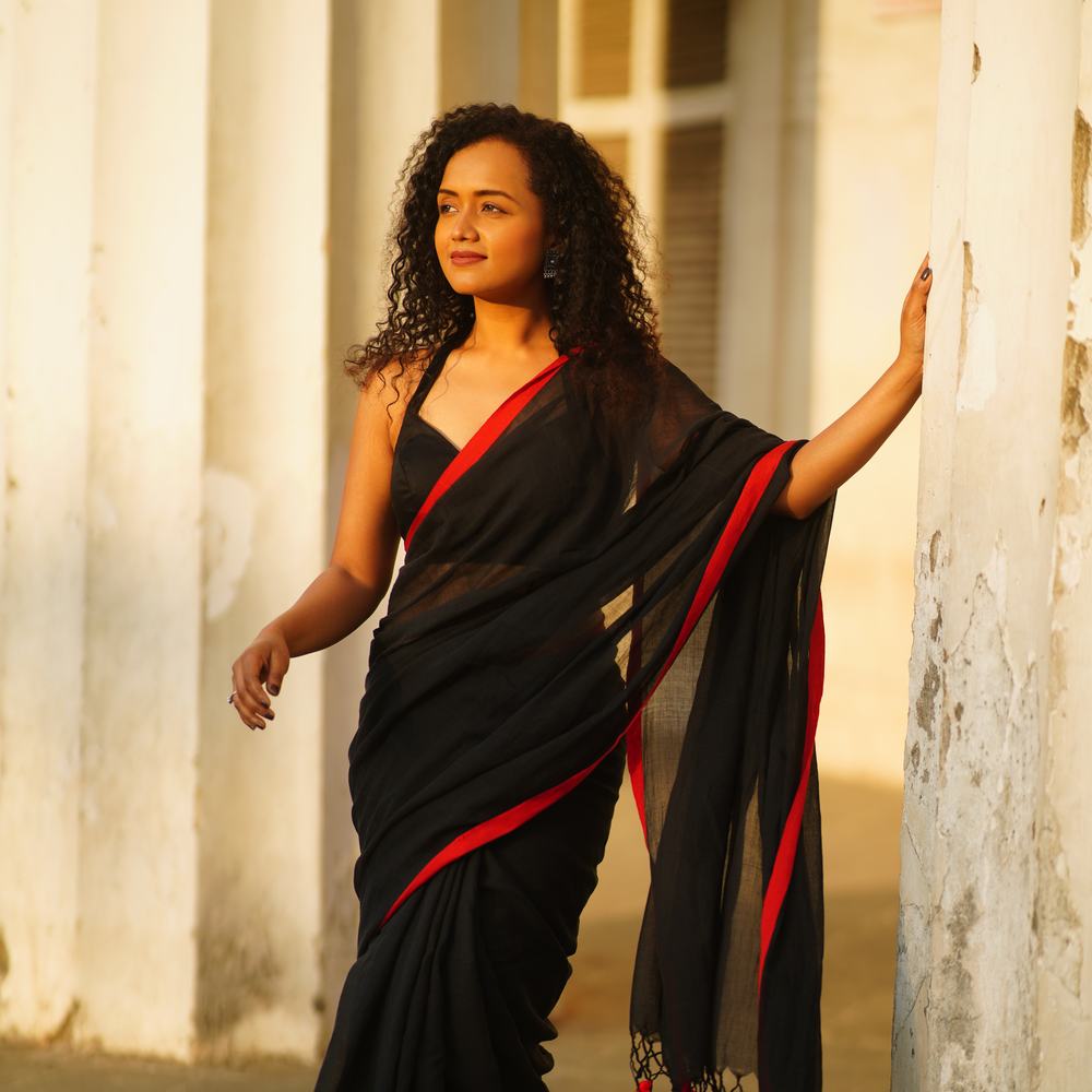 Pranali standing between pale columns in warm evening light, wearing a black sari with a red border.